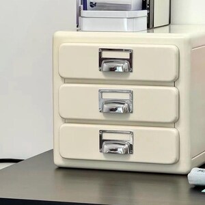 May include: A cream-colored, three-drawer cabinet with chrome handles. The cabinet is sitting on a black desk. The drawers are rectangular and have a simple, modern design. The handles are rectangular and have a shiny, reflective surface.