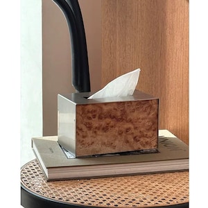 May include: A tissue box with a stainless steel top and a wooden burl veneer front. The box is rectangular and sits on a stack of books. White tissues are visible. The box is on a woven chair.