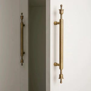 Peut inclure: Une paire de poignées de placard verticales avec une finition en laiton brossé. Chaque poignée présente une barre centrale cylindrique avec des embouts décoratifs en haut et en bas, fixée à une porte de placard blanche par deux supports.