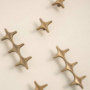 May include: A collection of five brass-toned metal hardware pieces. Two pieces are elongated with three connected star-shaped points, while three pieces are individual four-pointed star-shaped knobs with a weathered, antique finish.