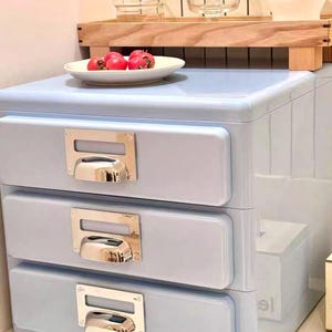 May include: A light blue three-drawer cabinet with silver metal handles. A white plate with red fruit sits on top. The cabinet is in a bathroom setting, with a wooden shelf holding glasses and a white cup.