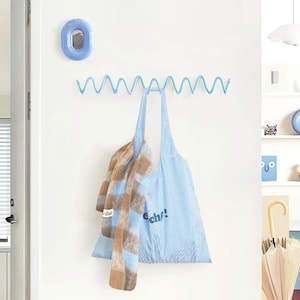 Könnte beinhalten: Eine hellblaue Wandgarderobe mit einem durchgehenden Zickzack- oder Wellendesign. Eine hellblaue gemusterte Tragetasche und ein braun-blauer Schal mit Karomuster hängen an der Garderobe. Darüber befindet sich ein kleiner, ovaler, hellblauer Wandspiegel.