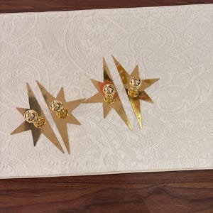 K&ouml;nnte beinhalten: Vier sternf&ouml;rmige Wandleuchten oder Zierplatten aus goldfarbenem Metall, jeweils mit einem zentralen kugelf&ouml;rmigen Knopf. Die sternf&ouml;rmigen R&uuml;ckplatten haben scharfe, l&auml;ngliche Spitzen und eine reflektierende, polierte Oberfl&auml;che.