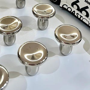 Puede incluir: Una colecci&oacute;n de pomos de metal plateado pulido con forma de seta y superficies lisas y reflectantes. Cada pomo presenta una tapa redondeada y una base cil&iacute;ndrica. Se ve un coche de juguete blanco con el texto BUZZRAW Mot 66 COASTCY.