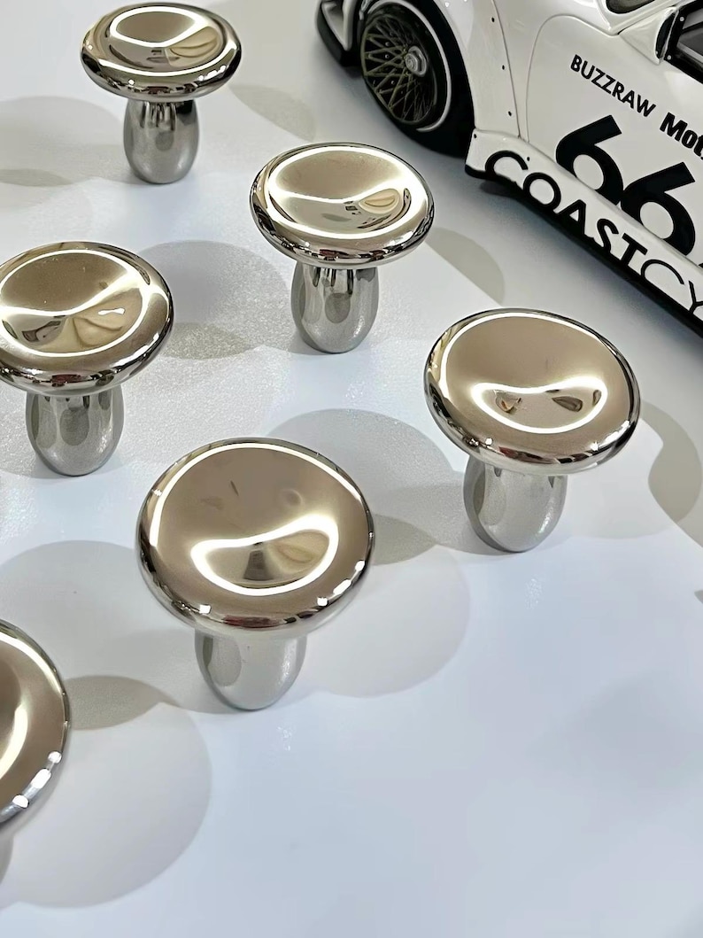 Puede incluir: Una colecci&oacute;n de pomos de metal plateado pulido con forma de seta y superficies lisas y reflectantes. Cada pomo presenta una tapa redondeada y una base cil&iacute;ndrica. Se ve un coche de juguete blanco con el texto BUZZRAW Mot 66 COASTCY.
