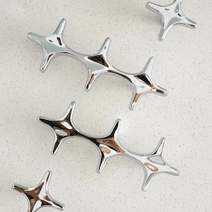 May include: A collection of shiny, chrome-finished starburst-shaped cabinet pulls and knobs. The hardware has a reflective surface, with a modern, minimalist design. The pulls are elongated, while the knobs are smaller and more compact.