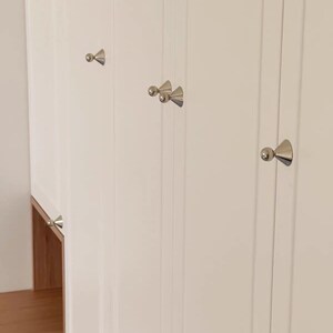 Puede incluir: Un conjunto de puertas de armario blancas con paneles verticales y pomos de metal plateado pulido. Cada pomo tiene una base c&oacute;nica rematada con una peque&ntilde;a esfera. Las puertas del armario son lisas y brillantes, con un estante de madera clara.