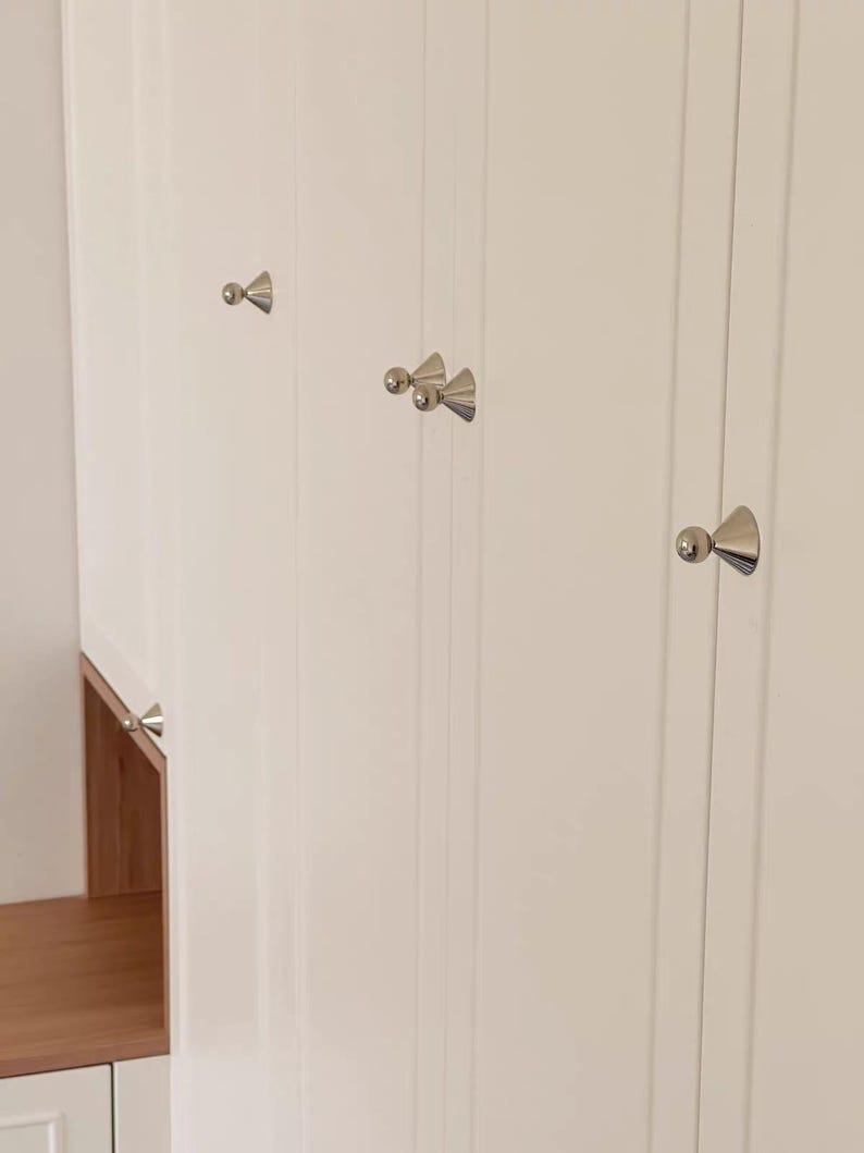 Puede incluir: Un conjunto de puertas de armario blancas con paneles verticales y pomos de metal plateado pulido. Cada pomo tiene una base c&oacute;nica rematada con una peque&ntilde;a esfera. Las puertas del armario son lisas y brillantes, con un estante de madera clara.