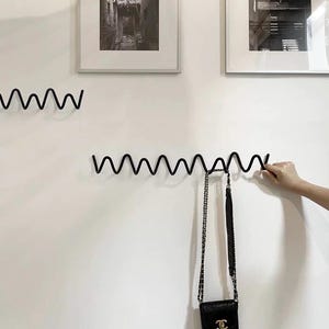 May include: A black, wavy metal wall hook with a small black purse hanging from it. The purse has a gold chain strap. Two framed black and white photos hang above the hook on a white wall.