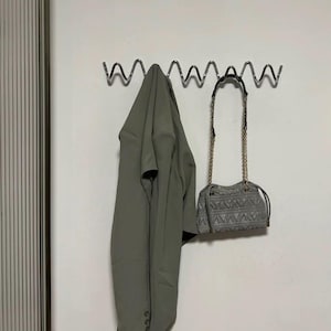 May include: A sage green blazer hangs on a wavy metal wall hook next to a gray quilted handbag with a gold chain strap. The blazer is buttoned and has a matching belt. The handbag has a geometric pattern.