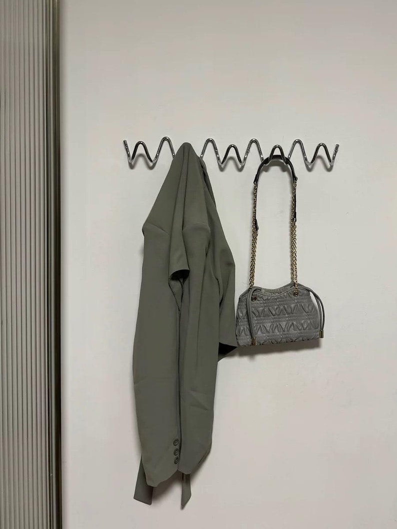 May include: A sage green blazer hangs on a wavy metal wall hook next to a gray quilted handbag with a gold chain strap. The blazer is buttoned and has a matching belt. The handbag has a geometric pattern.