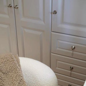Puede incluir: Armarios blancos con puertas de panel empotrado y tres cajones apilados con pomos plateados. Se ve una parte de una silla redondeada de tela texturizada color crema en primer plano, parcialmente cubierta por una manta de estilo sherpa color canela.