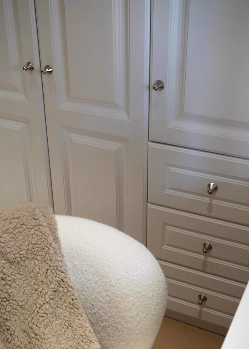Puede incluir: Armarios blancos con puertas de panel empotrado y tres cajones apilados con pomos plateados. Se ve una parte de una silla redondeada de tela texturizada color crema en primer plano, parcialmente cubierta por una manta de estilo sherpa color canela.