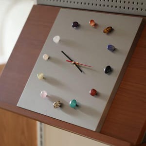 May include: A square wall clock with a gray face featuring twelve polished, multi-colored gemstone hour markers. The clock has two black hands and a thin red second hand. It is mounted on a wooden stand with a perforated gray metal panel above.