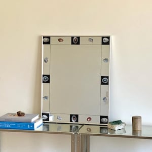 Könnte beinhalten: Ein rechteckiger Wandspiegel mit einem silbernen Rahmen, der mit abwechselnden schwarzen Quadraten und kleinen, detaillierten Illustrationen verschiedener Muscheln verziert ist. Die Spiegelfläche ist klar und reflektierend.