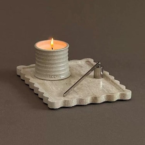 May include: A lit candle in a gray, ribbed ceramic container with the word "LOEWE" printed on the side. The candle sits on a decorative, scalloped-edge tray with a silver metal wick trimmer.