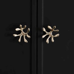 May include: Two metallic, gold-toned cabinet knobs with an irregular, organic, branch-like shape. The knobs are mounted on a smooth, matte black surface, positioned side-by-side with a vertical seam running between them.