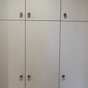 Puede incluir: Un conjunto de puertas de armario blancas dispuestas en cuadr&iacute;cula, con finas l&iacute;neas verticales y horizontales que separan cada panel. Cada puerta cuenta con un peque&ntilde;o pomo met&aacute;lico plateado y redondo centrado en el borde.