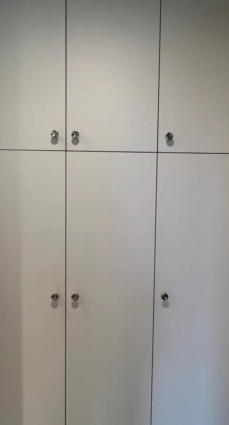 Puede incluir: Un conjunto de puertas de armario blancas dispuestas en cuadr&iacute;cula, con finas l&iacute;neas verticales y horizontales que separan cada panel. Cada puerta cuenta con un peque&ntilde;o pomo met&aacute;lico plateado y redondo centrado en el borde.