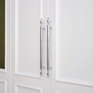 May include: Two vertical polished chrome door handles mounted on white paneled double doors. Each handle features a long cylindrical bar with decorative finials at the top and bottom, secured to the door surface by two small circular mounting brackets.
