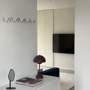 May include: A modern interior scene featuring a white wall with a decorative chrome hook rack and a white clock. A white dresser holds a small mirror, a magazine, and a burgundy lamp. A television is mounted on the wall in the background.