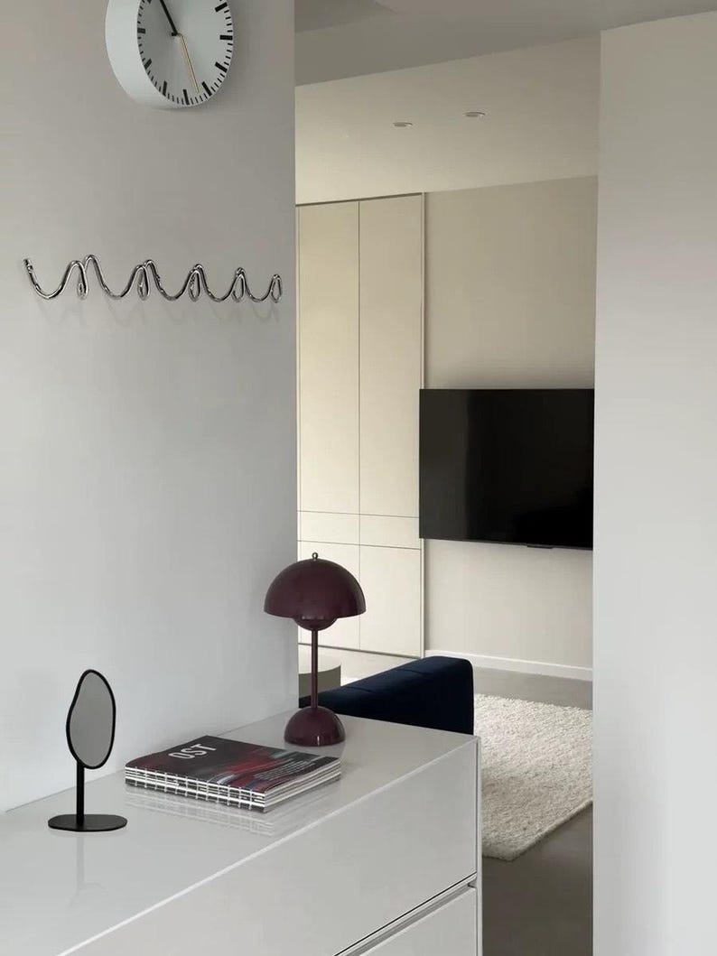 May include: A modern interior scene featuring a white wall with a decorative chrome hook rack and a white clock. A white dresser holds a small mirror, a magazine, and a burgundy lamp. A television is mounted on the wall in the background.