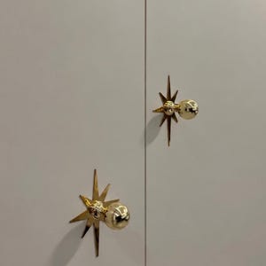 May include: Two gold-colored metal cabinet knobs mounted on a light gray double-door surface. Each knob features a spherical handle centered on a flat, multi-pointed starburst backplate with sharp, radiating rays extending outward.