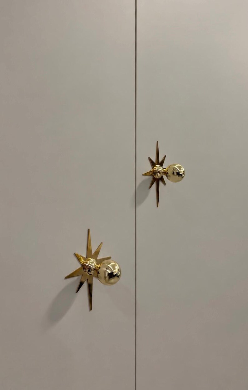 May include: Two gold-colored metal cabinet knobs mounted on a light gray double-door surface. Each knob features a spherical handle centered on a flat, multi-pointed starburst backplate with sharp, radiating rays extending outward.