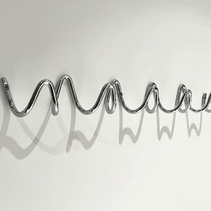 May include: A chrome-finished wall-mounted coat rack in a cursive "maaas" design. The rack is made of curved metal tubing and casts a shadow on the white wall. The design is modern and minimalist.