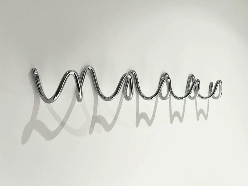 May include: A chrome-finished wall-mounted coat rack in a cursive "maaas" design. The rack is made of curved metal tubing and casts a shadow on the white wall. The design is modern and minimalist.