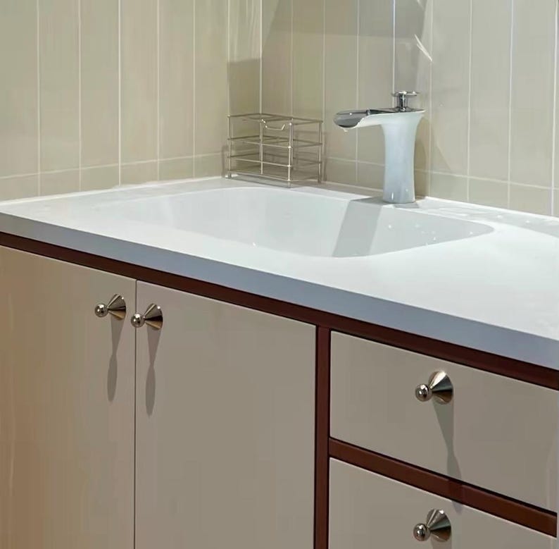 Puede incluir: Un lavabo blanco con grifo cromado, integrado en un mueble de ba&ntilde;o con puertas y cajones de color beige claro con peque&ntilde;os pomos plateados c&oacute;nicos. Un peque&ntilde;o organizador de alambre met&aacute;lico se encuentra sobre la encimera blanca.