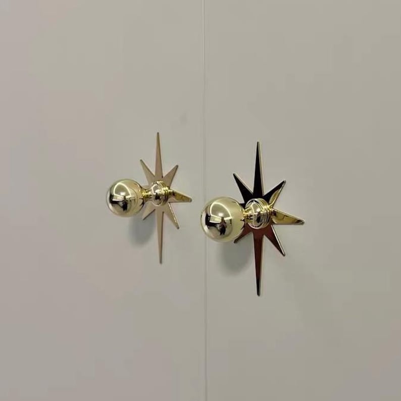 May include: Two polished gold-tone cabinet knobs mounted on a plain white surface. Each knob features a spherical handle centered on a flat, multi-pointed star-shaped backplate with elongated rays extending outward in a symmetrical design.