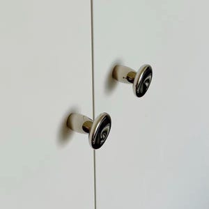May include: Two polished chrome cabinet knobs with a rounded, slightly flattened shape, mounted on a smooth white cabinet door surface. The metallic finish reflects light, creating a bright, reflective appearance on the hardware.