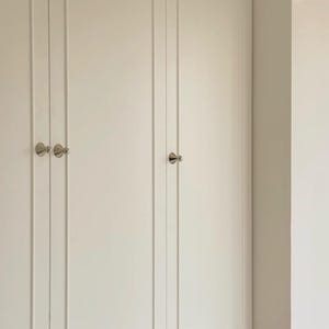 Puede incluir: Un conjunto de puertas de armario en color blanco roto con paneles verticales empotrados y peque&ntilde;os pomos redondos de metal plateado. Las puertas est&aacute;n montadas al ras y muestran un acabado limpio y uniforme con herrajes bien colocados.