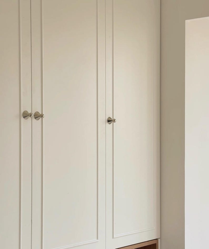 Puede incluir: Un conjunto de puertas de armario en color blanco roto con paneles verticales empotrados y peque&ntilde;os pomos redondos de metal plateado. Las puertas est&aacute;n montadas al ras y muestran un acabado limpio y uniforme con herrajes bien colocados.