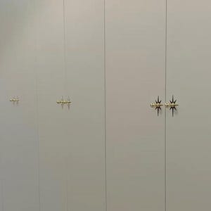 May include: A series of tall, light gray cabinet doors featuring gold-toned, starburst-shaped metal hardware. The handles are positioned in pairs on the vertical seams of the doors, creating a repeating pattern across the flat, smooth surface.