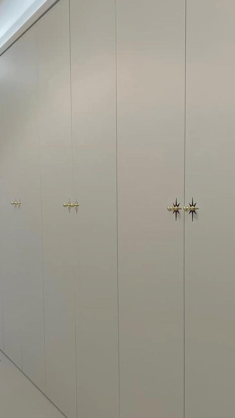 May include: A series of tall, light gray cabinet doors featuring gold-toned, starburst-shaped metal hardware. The handles are positioned in pairs on the vertical seams of the doors, creating a repeating pattern across the flat, smooth surface.