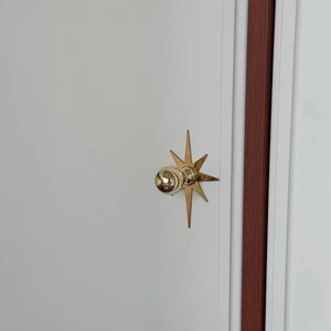 May include: A polished gold-tone metal door knob featuring a spherical handle mounted on a decorative starburst-shaped backplate. The hardware is attached to a smooth white door panel with a vertical dark brown trim detail visible on the right side.
