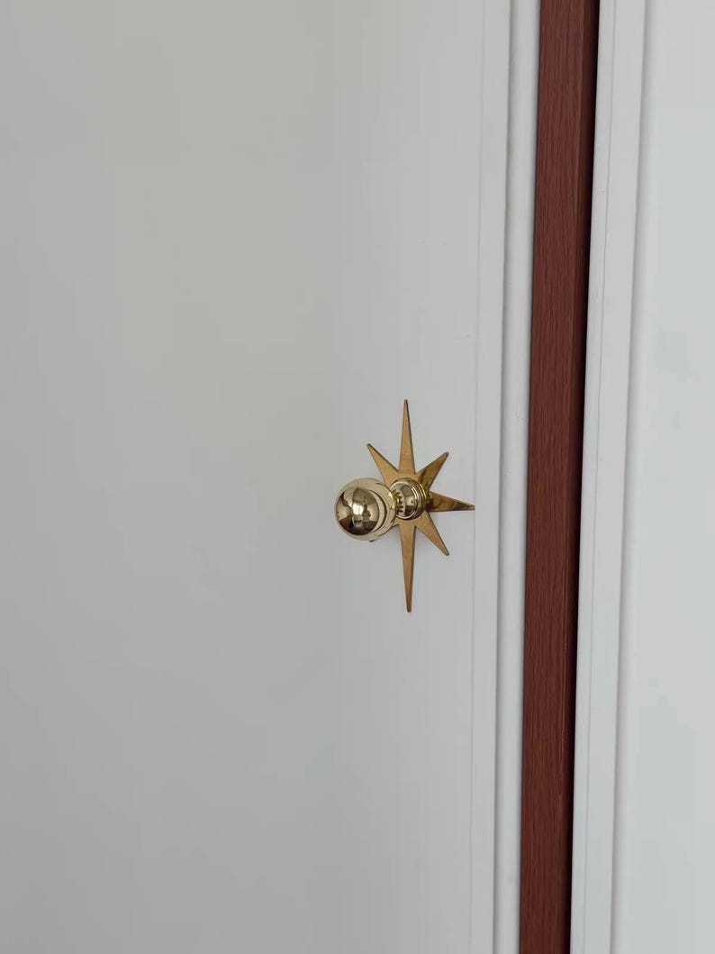 May include: A polished gold-tone metal door knob featuring a spherical handle mounted on a decorative starburst-shaped backplate. The hardware is attached to a smooth white door panel with a vertical dark brown trim detail visible on the right side.