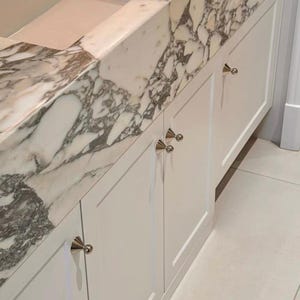 Puede incluir: Un mueble de ba&ntilde;o con puertas blancas de estilo shaker y pomos c&oacute;nicos de plata pulida. La encimera es de m&aacute;rmol blanco con vetas grises prominentes, integrada con un lavabo rectangular y un salpicadero a juego en el mismo material.