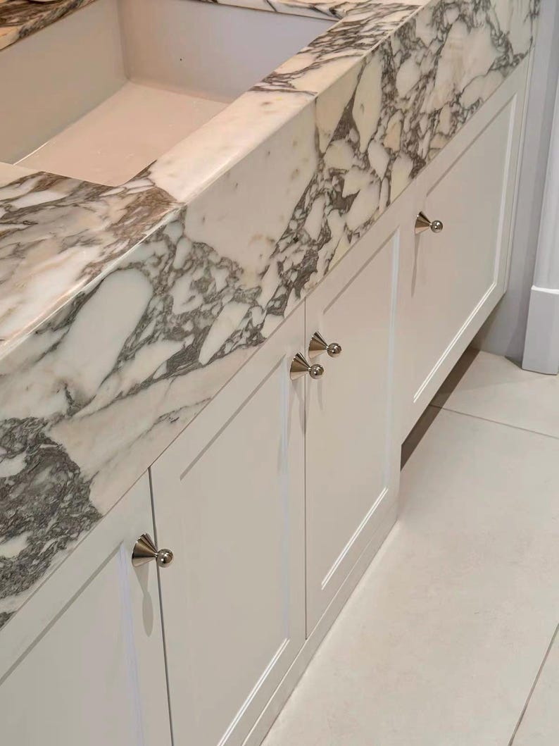 Puede incluir: Un mueble de ba&ntilde;o con puertas blancas de estilo shaker y pomos c&oacute;nicos de plata pulida. La encimera es de m&aacute;rmol blanco con vetas grises prominentes, integrada con un lavabo rectangular y un salpicadero a juego en el mismo material.