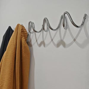 May include: A chrome-finished wall-mounted coat rack with a unique, wavy design. The rack is mounted on a white wall, with a black and a mustard-colored garment hanging from the hooks. The design is modern and minimalist.