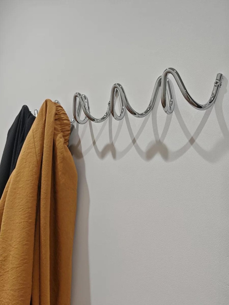 May include: A chrome-finished wall-mounted coat rack with a unique, wavy design. The rack is mounted on a white wall, with a black and a mustard-colored garment hanging from the hooks. The design is modern and minimalist.