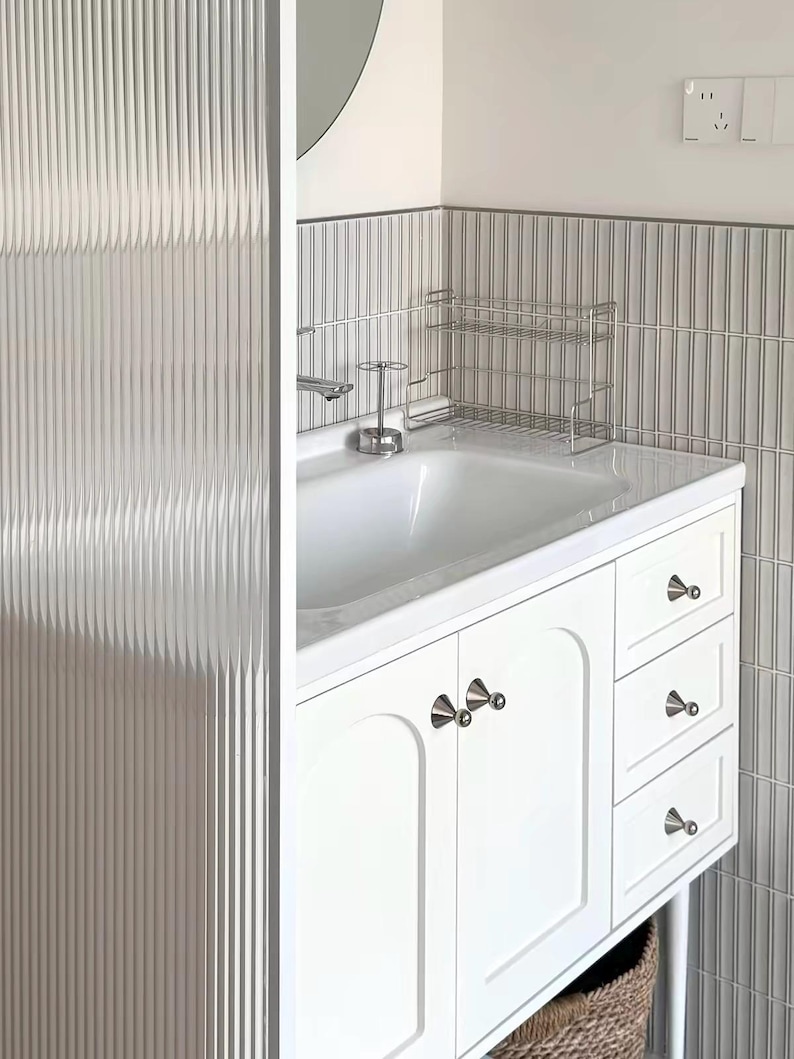 Puede incluir: Un mueble de ba&ntilde;o blanco con dos puertas y tres cajones con pomos c&oacute;nicos plateados. El tocador incluye un lavabo blanco, un grifo cromado, un peque&ntilde;o estante de alambre met&aacute;lico y una mampara de privacidad de vidrio estriado vertical.