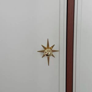 May include: A gold-colored starburst-shaped cabinet knob with a central spherical protrusion, mounted on a flat white door panel next to a vertical dark wood-toned trim piece. The hardware features eight pointed rays extending outward.