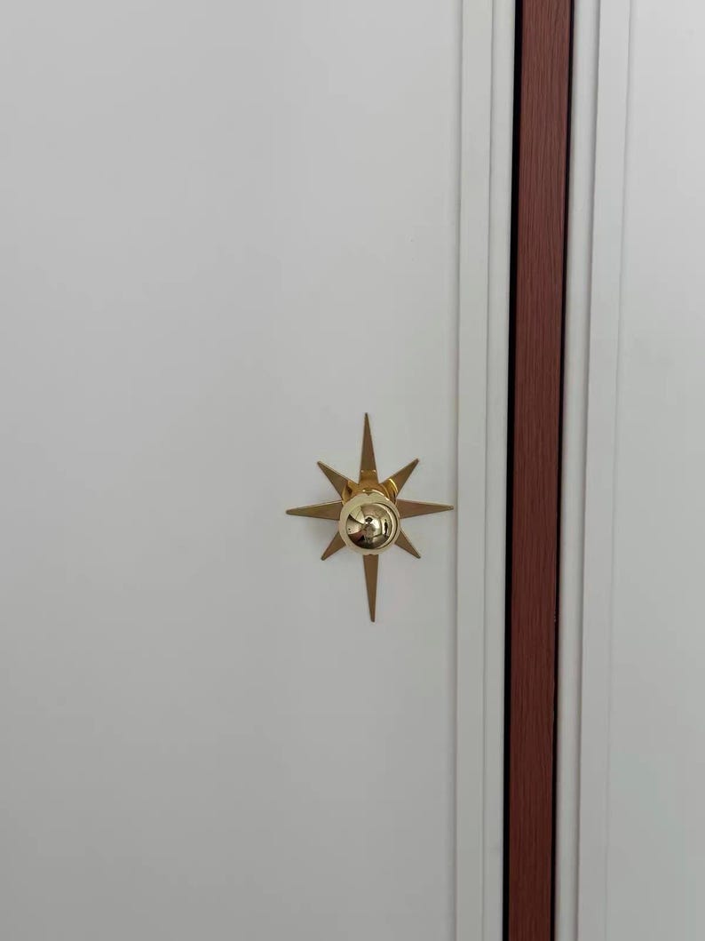 May include: A gold-colored starburst-shaped cabinet knob with a central spherical protrusion, mounted on a flat white door panel next to a vertical dark wood-toned trim piece. The hardware features eight pointed rays extending outward.