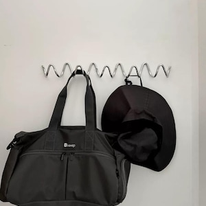 May include: A dark gray duffel bag and a black hat hanging on a silver metal wall rack. The duffel bag has two zippered pockets and the word "keep" printed on it. The hat is a wide-brimmed style.