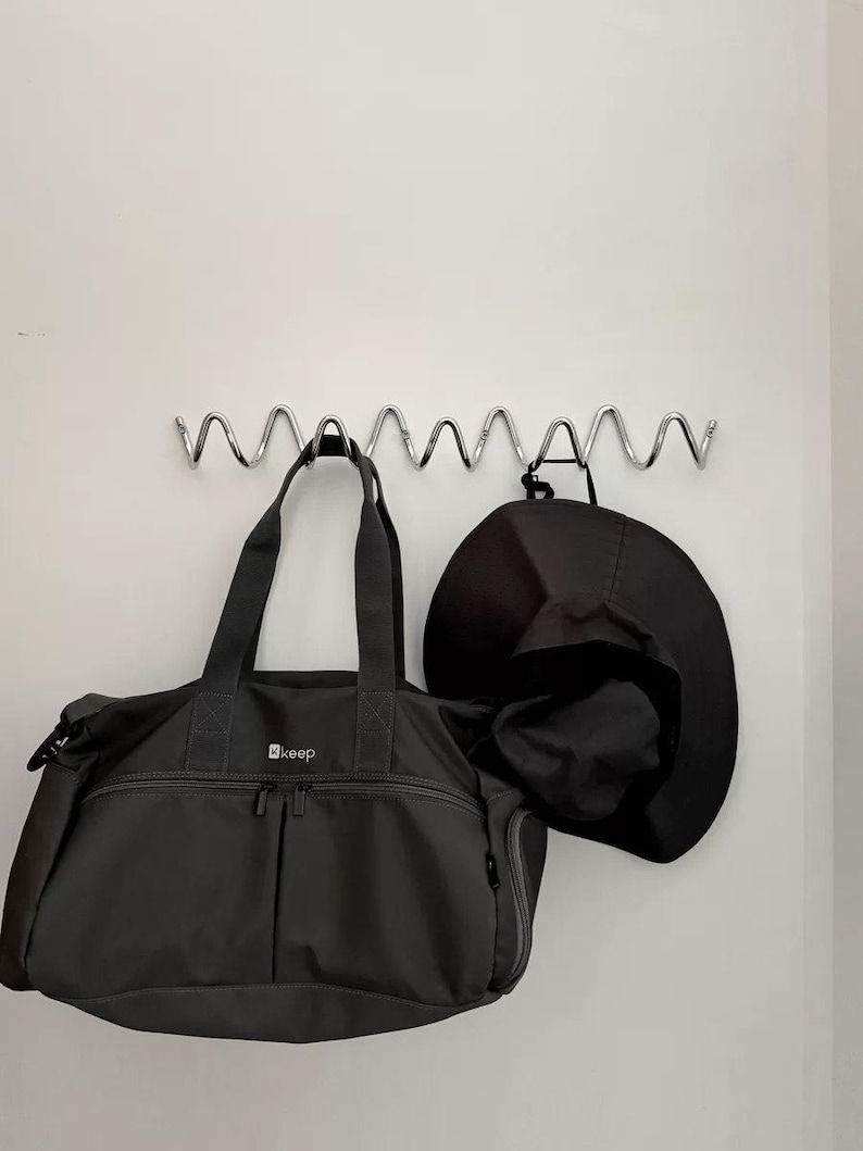 May include: A dark gray duffel bag and a black hat hanging on a silver metal wall rack. The duffel bag has two zippered pockets and the word "keep" printed on it. The hat is a wide-brimmed style.