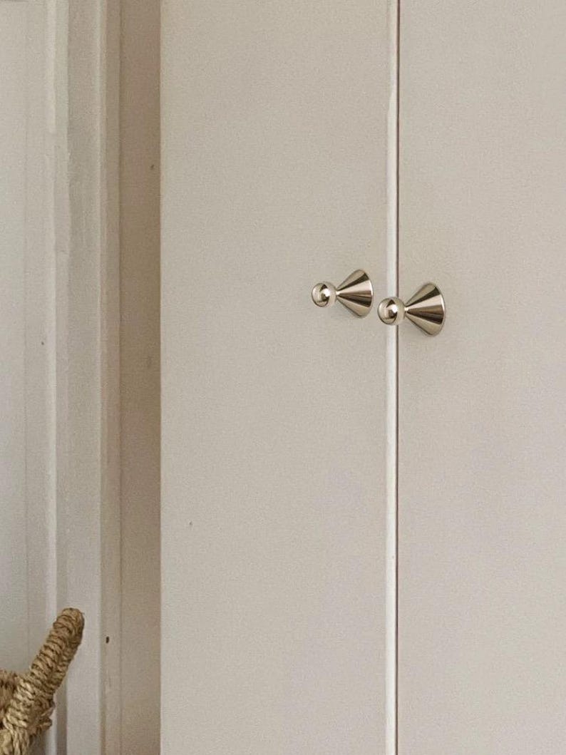 May include: Two polished silver-toned metal cupboard knobs with a conical base and a spherical top, mounted on a pair of light-coloured vertical cupboard doors that meet in the centre with a thin vertical gap between them.