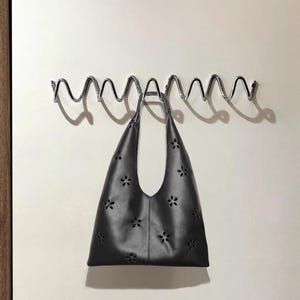 Può includere: Una borsa tote nera con un design a fiori traforati è appesa a un attaccapanni ondulato in metallo cromato. La borsa ha un manico singolo e un'ampia apertura. Lo sfondo è una parete bianca sporca.